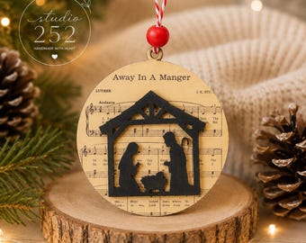 Könnte beinhalten: Ein runder Weihnachtsanhänger aus Holz mit einer schwarzen Silhouette einer Krippenszene in einem Stall. Der Hintergrund zeigt gedruckte Notenblätter mit dem Text Away In A Manger. Er hängt an einer rot-weißen Schnur.