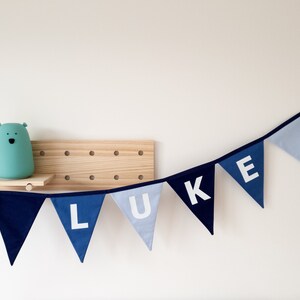 May include: A blue fabric banner spelling "LUKE" in white letters. The pennants are in shades of blue, with a dark blue border. A light blue bear-shaped lamp sits on a wooden shelf.