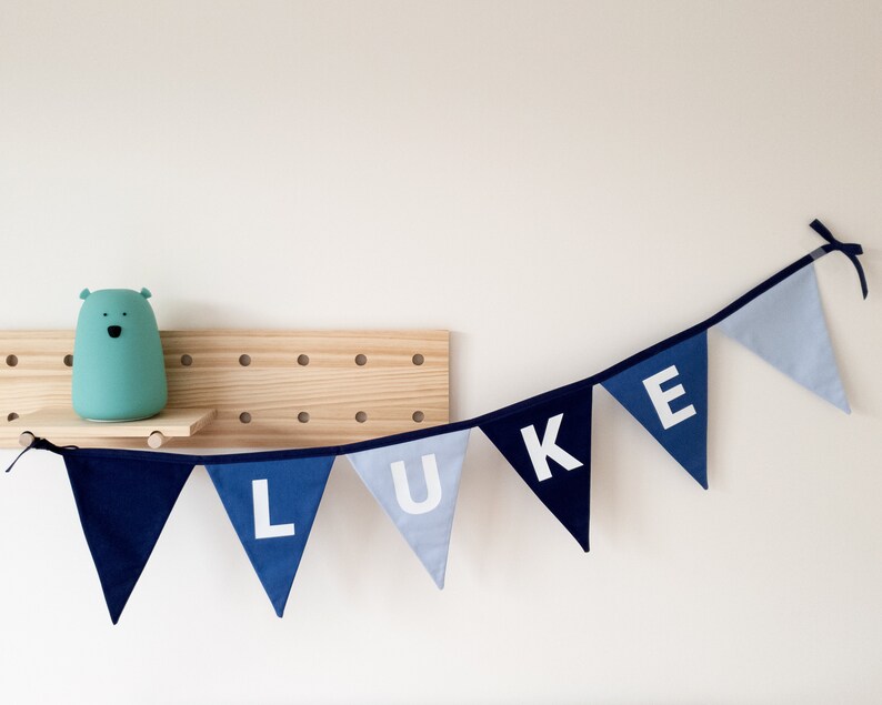 May include: A blue fabric banner spelling "LUKE" in white letters. The pennants are in shades of blue, with a dark blue border. A light blue bear-shaped lamp sits on a wooden shelf.