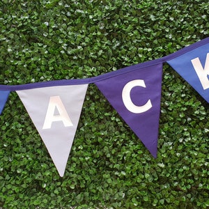 May include: A banner with triangular flags spelling out the name "JACK". The flags are in shades of blue and gray, with white letters. The banner is against a backdrop of green foliage.