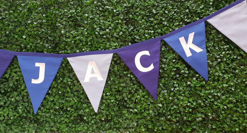 May include: A banner with triangular flags spelling out the name "JACK". The flags are in shades of blue and gray, with white letters. The banner is against a backdrop of green foliage.