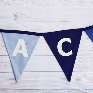 May include: A blue fabric banner spelling out the name "JACK" in white capital letters. The banner consists of triangular flags in shades of blue, strung together with a dark blue ribbon. The background is a light-colored wooden surface.