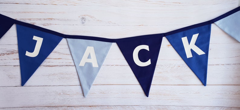 May include: A blue fabric banner spelling out the name "JACK" in white capital letters. The banner consists of triangular flags in shades of blue, strung together with a dark blue ribbon. The background is a light-colored wooden surface.
