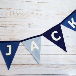 May include: A blue pennant banner spelling out the name "JACK" in white letters. The banner features alternating shades of blue triangles, including navy and light blue, strung together with a dark blue trim. The background is a white wooden surface.