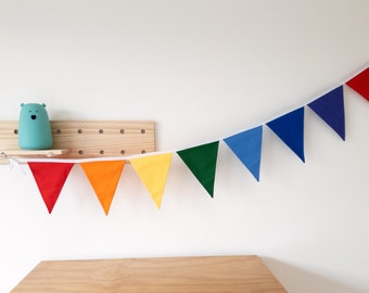 May include: A colorful rainbow banner with red, orange, yellow, green, blue, and purple triangular flags. The banner is strung on a white ribbon and hangs against a white wall. A wooden shelf with a blue bear-shaped lamp is visible.