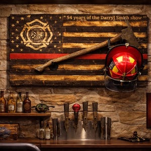 May include: A wooden wall art piece featuring a charred American flag design with a thin red line, a fire department emblem, and the text "54 years of the Darryl Smith show". A wooden axe and a red and yellow firefighter helmet are mounted on the flag.
