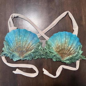 May include: A bikini top featuring two large, moulded seashell cups in a gradient of blue, green, and gold. The shells are adorned with intricate, raised scrollwork detailing at the base. The garment includes beige fabric straps that cross at the back.