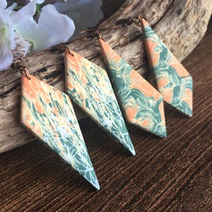 May include: Two pairs of diamond-shaped dangle earrings featuring a marbled pattern of peach, sage green, and white. Each earring is suspended from a dark metal hook, showcasing a smooth, flat surface with intricate, swirling color transitions.