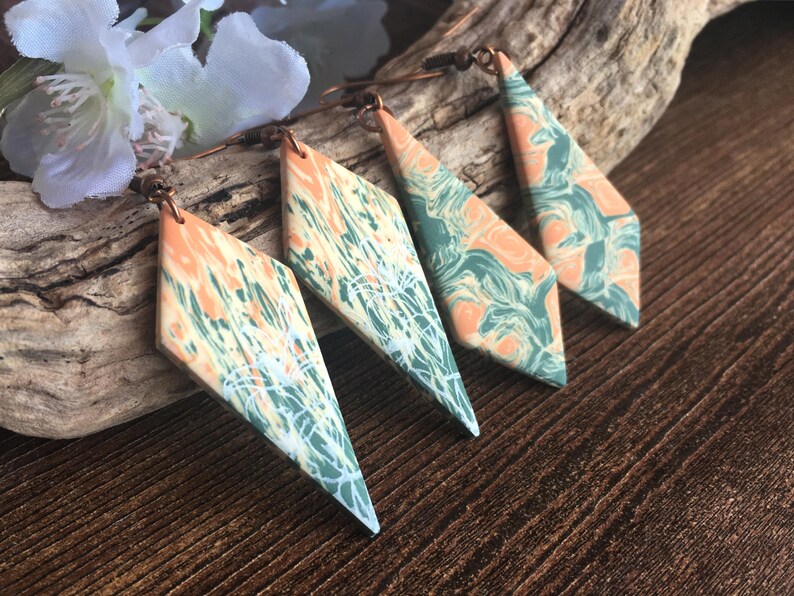 May include: Two pairs of diamond-shaped dangle earrings featuring a marbled pattern of peach, sage green, and white. Each earring is suspended from a dark metal hook, showcasing a smooth, flat surface with intricate, swirling color transitions.