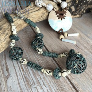 May include: Handmade necklace featuring a unique design with teal and cream-colored beads, including woven spheres and decorative elements. The necklace has a large, round pendant with a blue and white design and brown lacing. The necklace is displayed on a wooden surface.