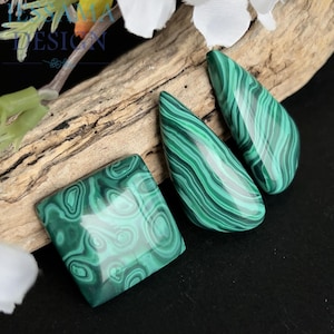 Puede incluir: Tres piedras preciosas de malaquita pulidas en tonos verdes. Una es cuadrada y dos tienen forma de lágrima. Las piedras preciosas tienen un patrón arremolinado y se exhiben sobre un trozo de madera flotante. La palabra "ESYAMA DESIGN" es visible.