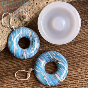 Könnte beinhalten: Ohrringe in Donutform mit blauem, weißem und braunem Marmormuster, zusammen mit einer weißen, kreisförmigen Form. Die Ohrringe haben silberfarbene Klappbügelverschlüsse. Die Ohrringe liegen auf einer Holzoberfläche.