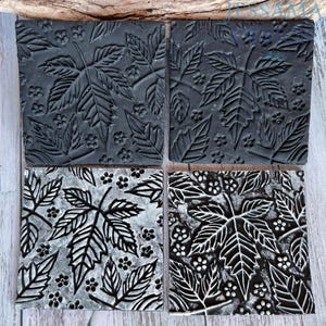 May include: Four square coasters featuring a raised relief pattern of maple leaves and small flowers. Two coasters are solid matte black, while the other two have a silver metallic finish applied over the black base to highlight the intricate texture.