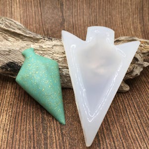 May include: A clear silicone mold shaped like an arrowhead, next to a green arrowhead pendant with gold glitter.
