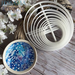 May include: A set of white circular nesting cutters with a cross-bar support structure and a small round bowl featuring a blue and white winter-themed pattern with snowflakes, swirling lines, and a stylised deer silhouette on the interior surface.