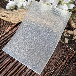 May include: A clear silicone mould with a pebble texture. The rectangular mould is angled, showing the rounded shapes. The background features wood and white flowers, suggesting a craft or DIY theme.