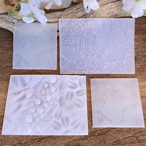May include: Four translucent resin coasters with embossed designs. One square coaster features delicate floral patterns, while another rectangular coaster showcases koi fish and aquatic plants. The other two coasters have a square and rectangular shape with other nature-inspired designs.