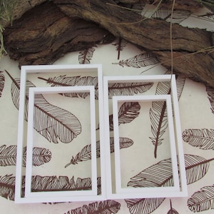 May include: Three white picture frames of different sizes, nested inside each other. The frames are on a background of brown feathers printed on a cream-coloured surface.