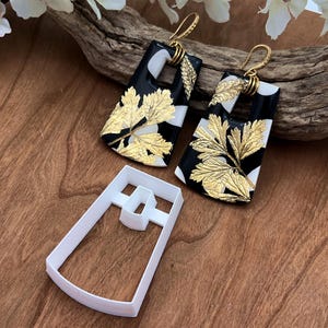 May include: A pair of black and white earrings with gold leaf accents. The earrings are shaped like a rectangle with a cutout in the center. The earrings are hanging from gold hooks.