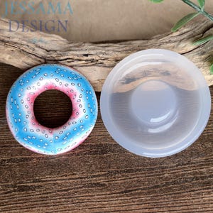 Pode incluir: Um molde de resina em forma de donut colorido ao lado de um donut de resina acabado. O donut tem uma cobertura azul com granulado branco e um centro rosa. O molde é transparente e circular. O texto "JESSAMA DESIGN" é visível.