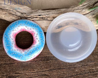 May include: A donut-shaped object with a blue and pink gradient surface featuring small white oval patterns, positioned next to a translucent, circular silicone mold. The text JESSAMA DESIGN is visible in the upper left corner of the image.