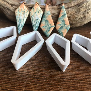 May include: Four diamond-shaped earrings featuring a marbled pattern of peach and sage green colors, accompanied by several white plastic diamond-shaped cutters. The earrings have copper-toned hardware at the top for attachment.