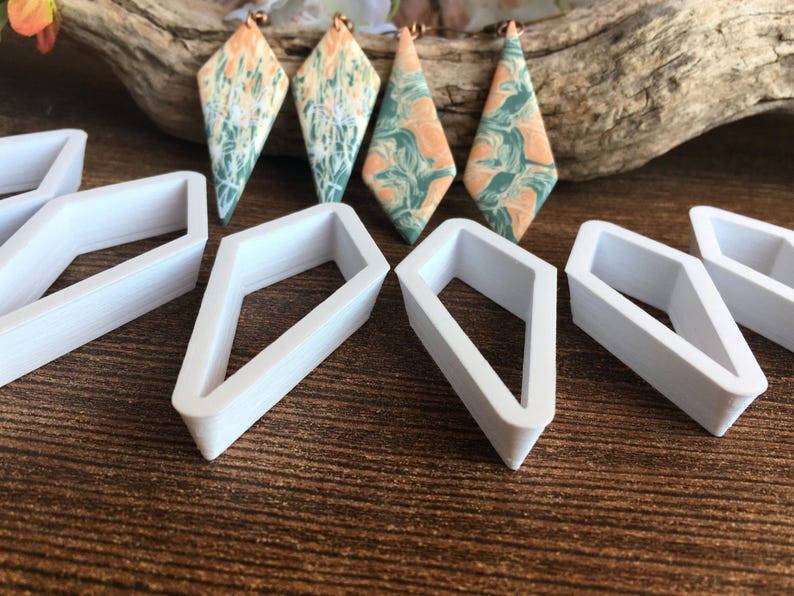 May include: Four diamond-shaped earrings featuring a marbled pattern of peach and sage green colors, accompanied by several white plastic diamond-shaped cutters. The earrings have copper-toned hardware at the top for attachment.