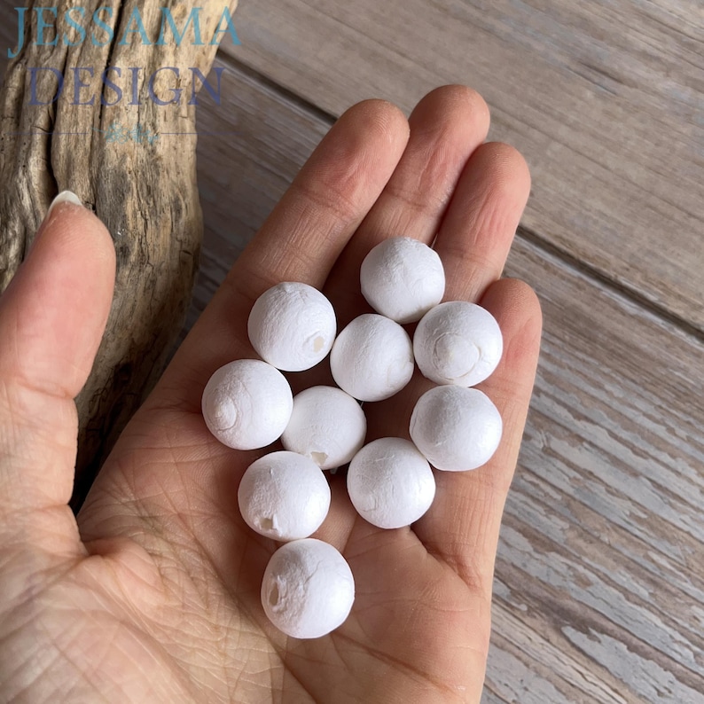 May include: A hand holds a collection of small, white, spherical beads. The beads have a textured surface and are arranged in a loose cluster. The background is a light-colored wooden surface.