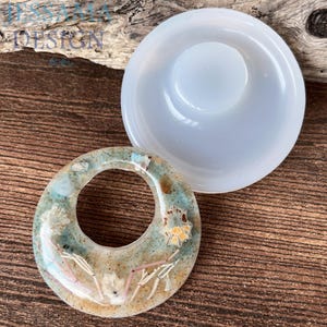 May include: A clear resin pendant with a circular design, featuring embedded dried flowers and other natural elements in shades of blue, beige, and white. A separate, light blue, circular mold is also visible. The pendant is approximately 5 cm in diameter.