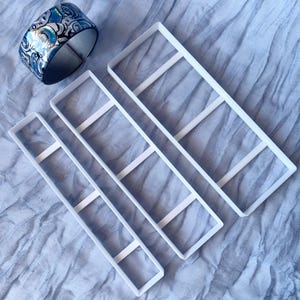 May include: A blue and white patterned bangle bracelet sits beside three white rectangular cookie cutters. The bracelet has a silver interior. The cookie cutters are rectangular with three horizontal bars.