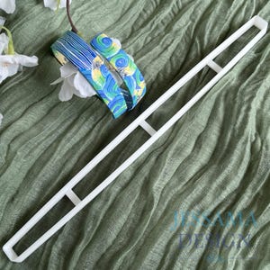 May include: A white, rectangular, open-frame tool with two crossbars, possibly a craft or design template, rests on green fabric. Two patterned headbands with blue, yellow, and green swirls and lines are also present, with white flowers.