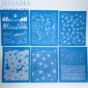 May include: Six rectangular blue screen printing stencils featuring white designs. The patterns include a mountain landscape, flying cranes, dandelion seeds, two variations of daisy flowers, and a grid texture. Each stencil is labeled JessamaDesign.