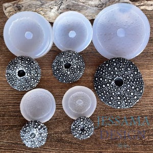 May include: An assortment of ear gauges and plugs in various sizes. The gauges are white and translucent, while the plugs are black with white speckles. The items are arranged on a wooden surface.