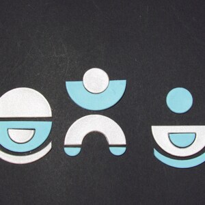 May include: Three abstract shapes made of white and light blue paper. The shapes are arranged in a row on a black background. The shapes are simple and geometric.