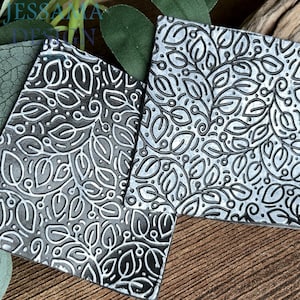 May include: Two square, silver metal coasters featuring a raised leaf and vine pattern. The coasters are positioned at an angle, highlighting the detailed design. The background includes green leaves and a wooden surface.