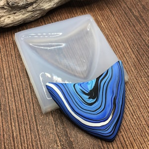 May include: A clear silicone mold for making resin jewelry. The mold is shaped like a triangle with rounded edges. A blue, black, and white resin piece is shown next to the mold.