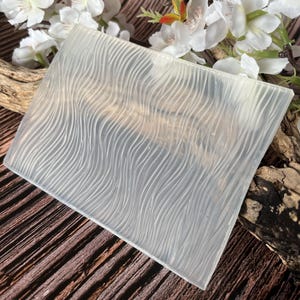 Op de afbeelding: Een heldere, vierkante mal met een golvend, getextureerd patroon. De mal is gemaakt van een doorschijnend materiaal en is ideaal voor knutselen. Witte bloemen en een houten tak staan op de achtergrond.