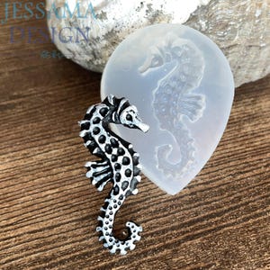 May include: A detailed black and white seahorse figurine next to a clear silicone seahorse mould. The seahorse is on a wooden surface. The figurine has a textured surface with spots and stripes.