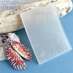 Peut inclure: Boucles d'oreilles artisanales avec un motif rouge, blanc et bleu, à côté d'un moule rectangulaire transparent et texturé. Les boucles d'oreilles sont en forme de goutte d'eau et présentent des motifs détaillés. Le moule a un motif de soleil. Le fond est bleu.