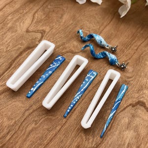 Pode incluir: Cinco moldes de plástico branco para fazer brincos, com quatro brincos com padrões azuis e prateados sobre uma superfície de madeira.