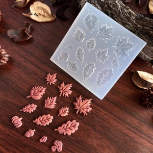 May include: A clear silicone mold with various leaf shapes, including maple and oak leaves, and small, reddish-brown leaf-shaped castings. The mold and castings are on a dark wooden surface, suggesting a fall or autumn theme.