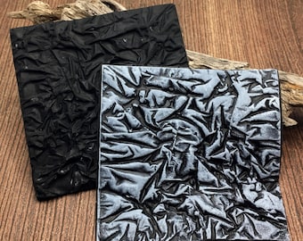 May include: Two square tiles with a highly textured, crinkled surface. One tile is solid matte black, while the other features a black base with a metallic silver finish applied to the raised ridges of the crumpled texture.