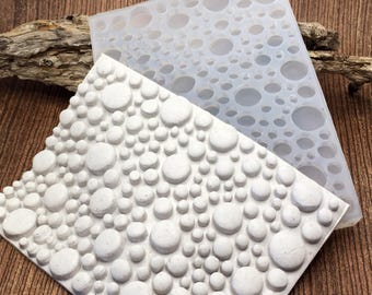 May include: Two silicone molds, one white and one translucent, with a pattern of varying-sized circles. The white mold has raised circles, while the translucent mold has recessed circles. These molds are likely for crafting or decorative purposes.