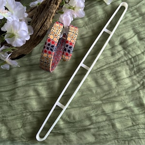 Op de afbeelding: Een witte, lange, rechthoekige polymeerklei snijder met drie dwarsbalken, naast twee polymeerklei armbanden met een geometrisch patroon in rood, geel en zwart. De armbanden liggen in de buurt van witte bloemen en een stuk hout op een groene stof.