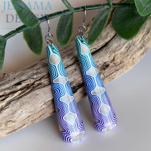 May include: A pair of long, tapered drop earrings featuring a gradient pattern of teal, blue, and purple wavy lines over a pearlescent white base. The earrings are suspended from silver-toned metal hooks and have a smooth, polished finish.