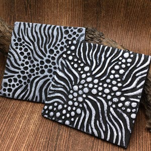 May include: Two black and silver square coasters with a raised design of wavy lines and circles.