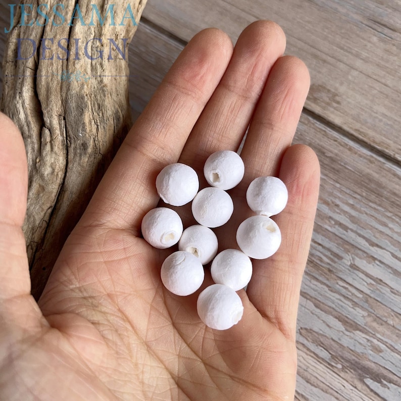 May include: A hand holding a collection of small, round, white beads. Each bead has a small hole. The beads are clustered together, and the hand is positioned against a light wooden surface. The image includes the text "JESSAMAIA DESIGN".