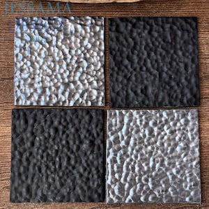 Könnte beinhalten: Vier quadratische Untersetzer in einer Gitteranordnung mit einer gehämmerten, genoppten Textur. Zwei Untersetzer haben eine metallisch silberne Oberfläche, während die anderen beiden eine mattschwarze Oberfläche aufweisen. Der Text JESSAMA ist sichtbar.