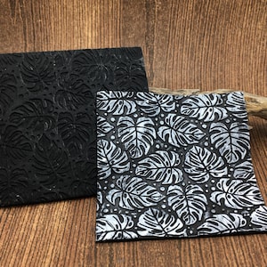 May include: Two square, textured coasters. One is solid black with a raised leaf pattern. The other is black with a silver leaf pattern. The coasters are made of a durable material and are designed to protect surfaces from heat and moisture.
