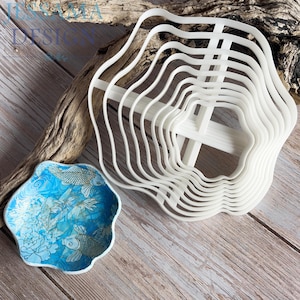 May include: A set of white, nested, wavy-edged plastic cutters arranged in a stack, accompanied by a small, scalloped-edge bowl featuring a blue and white graphic print of koi fish and floral patterns, with the text JESSAMIA DESIGN visible.